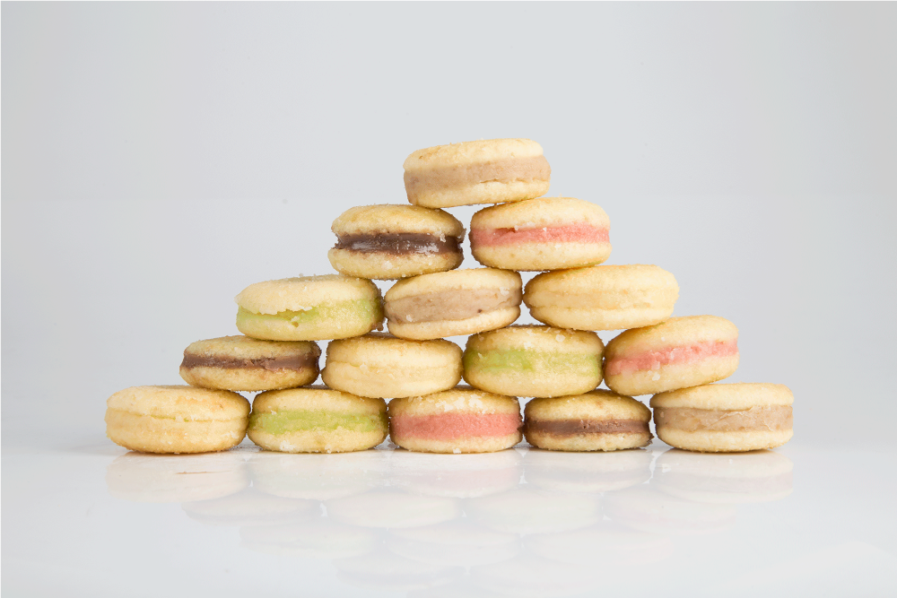 A imagem apresenta 15 unidades de bem casado da Zulmira Doces de diferentes sabores: tradicional de doce de leite, pistache, nozes, frutas vermelhas e chocolate que estão empilhados em formato de pirâmide